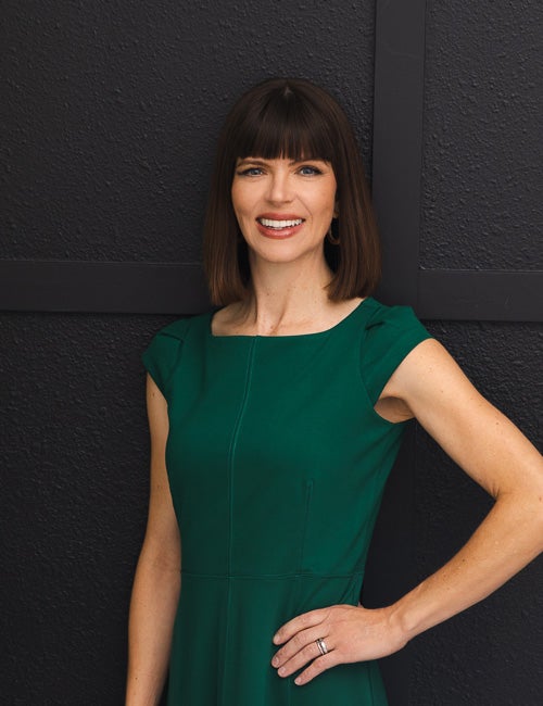 A headshot of a woman with dark hair wearing a green dress, smiling. She is posed with her hand on her hip and is looking directly at the camera. 