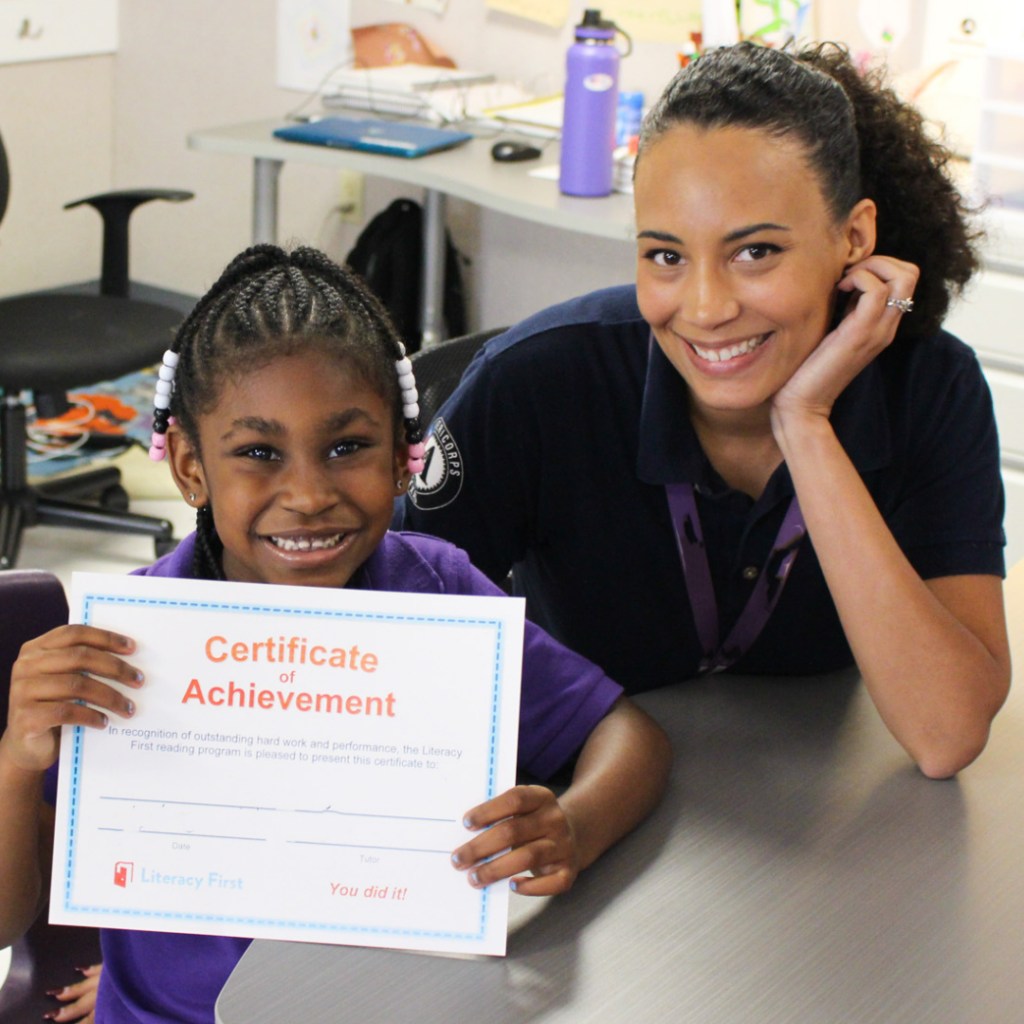Bri sits at a table, smiling and looking at the camera. A young student sits next to her, holding a Certificate of Achievement and also smiling at the camera.
