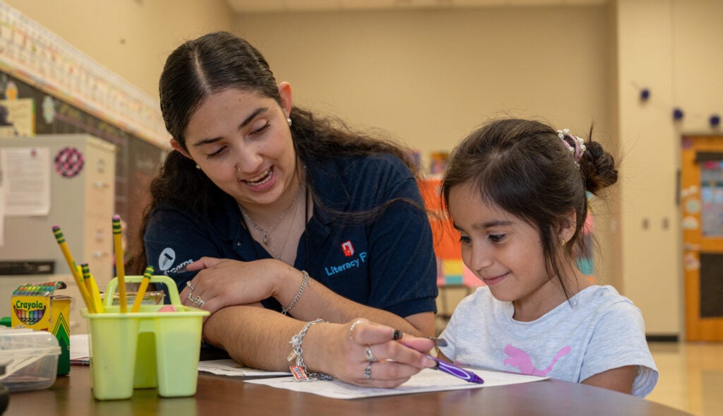 An adult and a child sit side by side, reading from a paper together. 