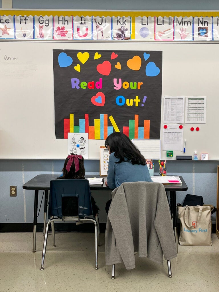 A woman sits next to a child at a desk, reading from a workbook. Their backs are to the camera and there is a poster on the wall that says "read your heart out."